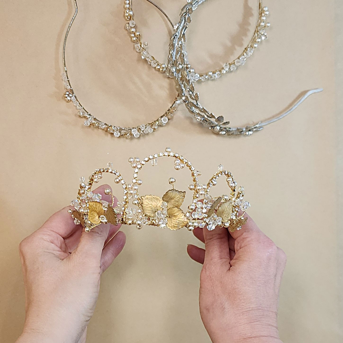 Hands holding a gold leaf and beaded tiara, with three embellished headbands arranged on the table behind it.