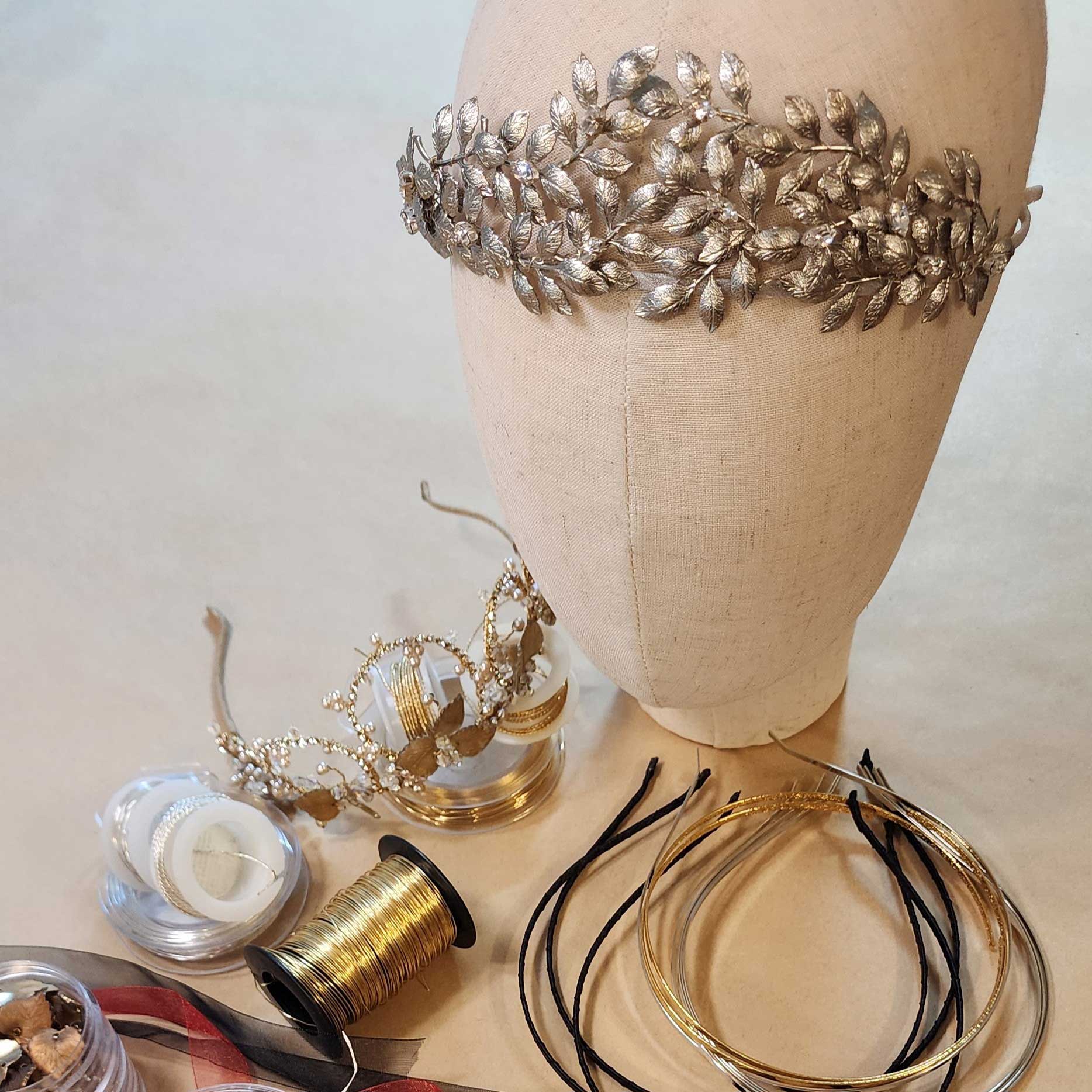 Metallic leaf headpiece on a fabric display head, with a gold tiara, wire spools and headband bases below.