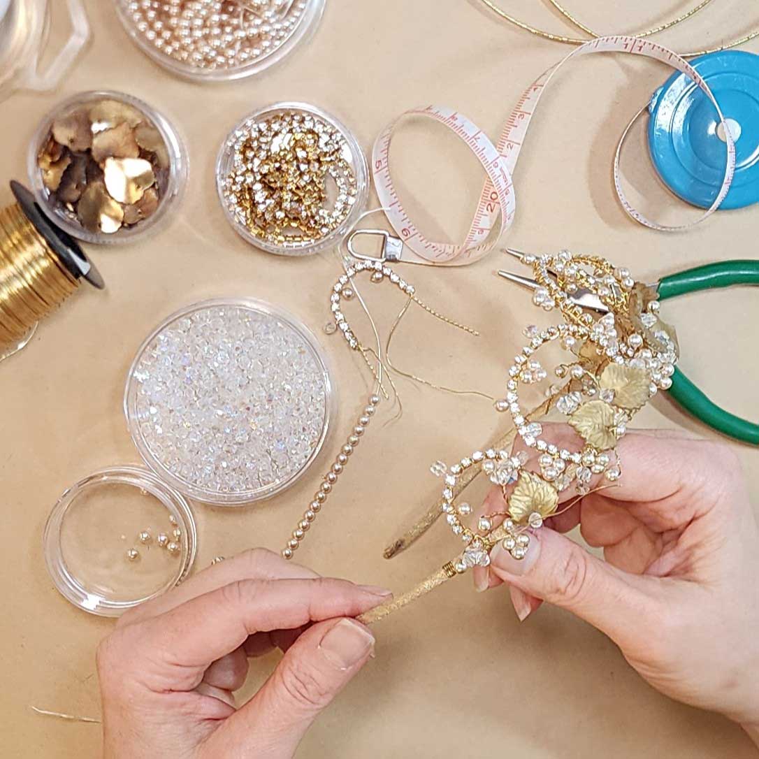Hands holding the wire-wrapped band of a gold leaf and beaded tiara at a worktable with beads and pliers.